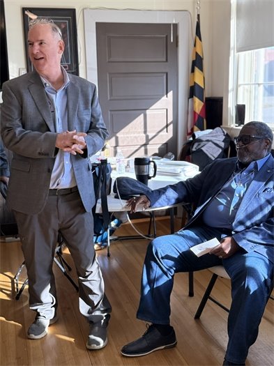 one man standing and talking and another sitting in a chair